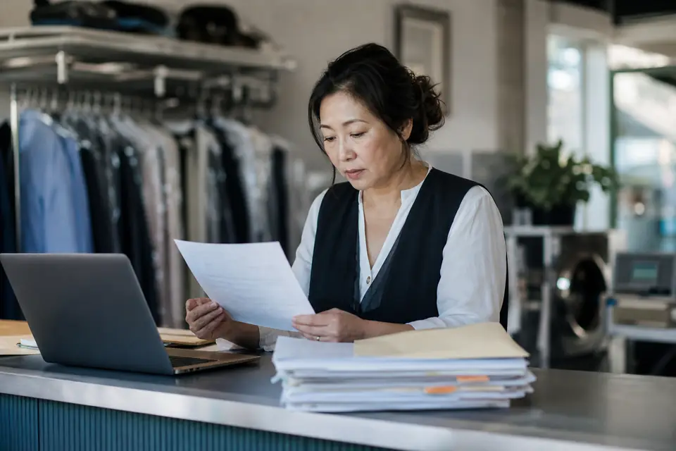 Small shop owner behind a counter reviewing a letter and payroll records