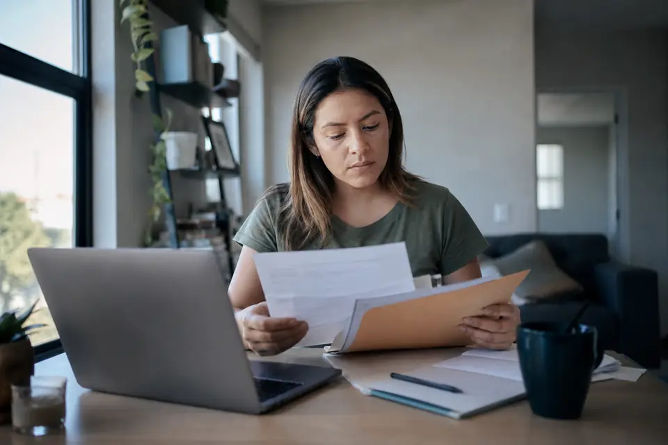 Woman at an apartment desk with a laptop and a folder of family papers