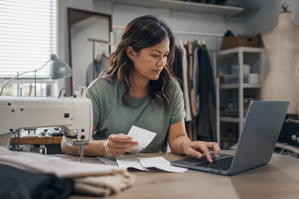 Self-employed tailor entering business receipts into a laptop in her home workshop